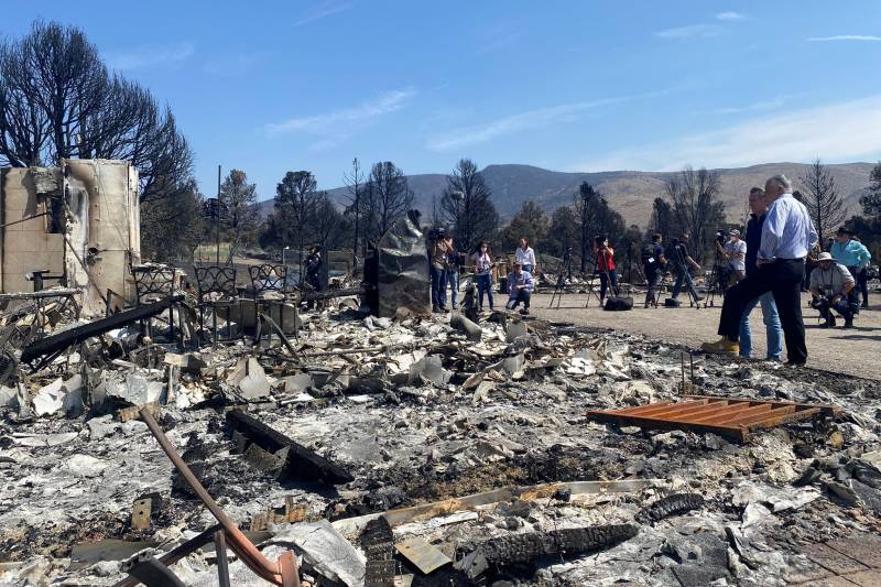 The image centers on the burnt remains of several homes. A fire has just passed by. To the right, two figures are visible observing the ruins. They are California Governor Gavin Newsom and Nevada Governor Steve Sisolak.