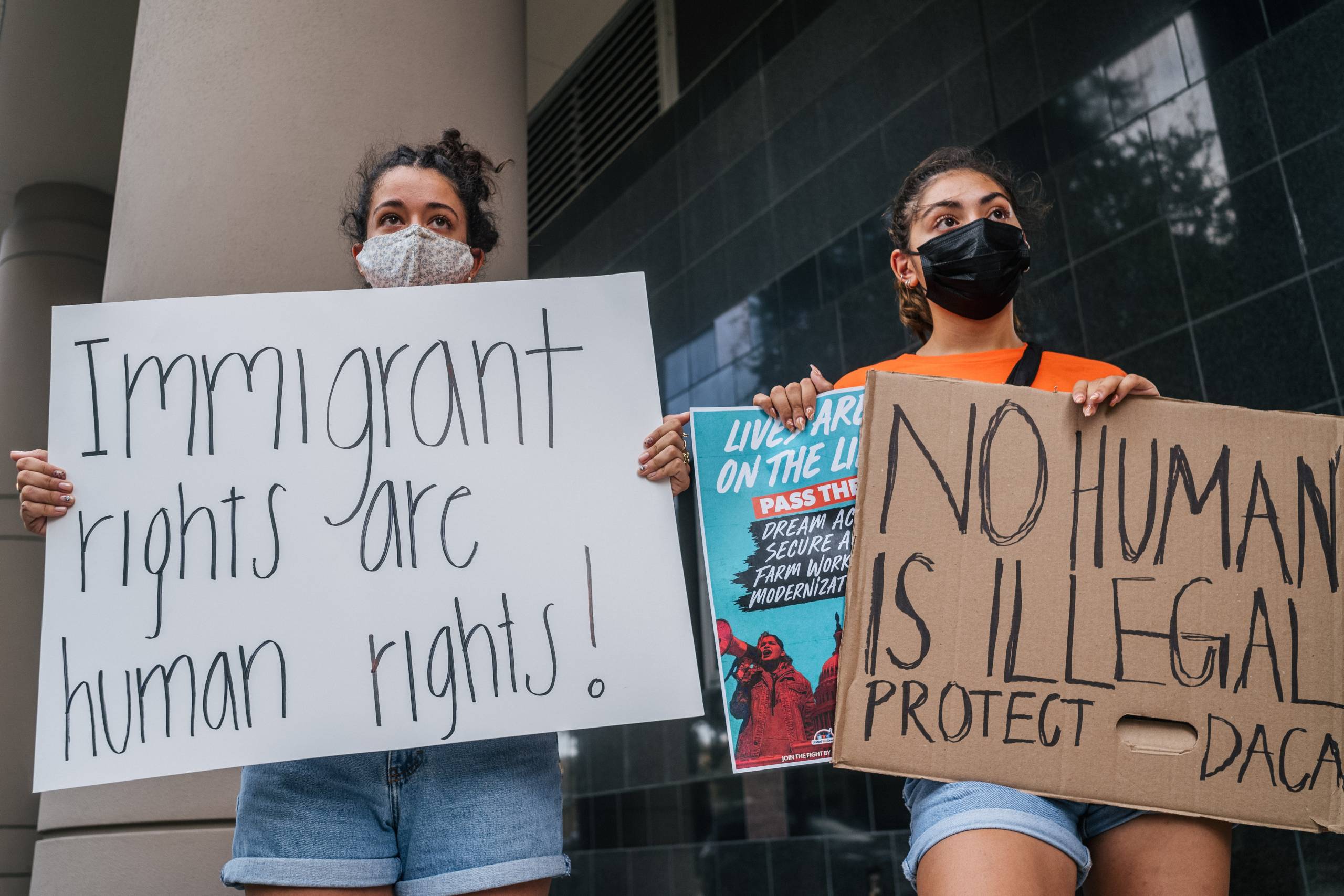 Two people wearing facemasks hold up signs that say, 'Immigrant rights are human rights!' and 'No human is illegal. Protect DACA.'