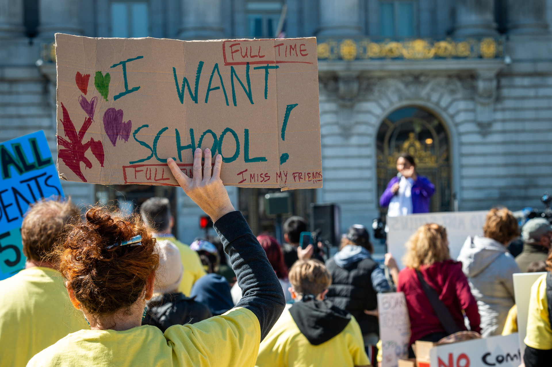 Protest sign saying "I want school! Full time. I miss my friends" in foreground of crowd before City Hall. Protestors in yellow T-shirts, Mayor Breed at podium in background.