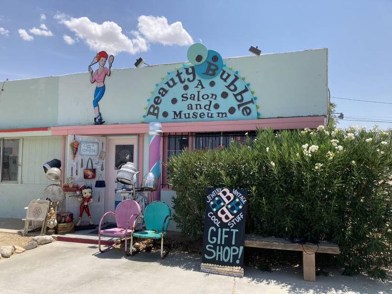 A blue-and-pink storefront sits in a desert with a blue sky above and green bushes out front.
