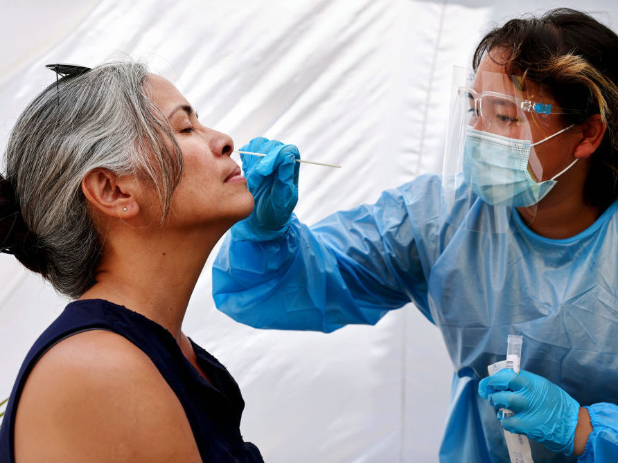 A medical assistant stands over a woman and takes a swab from her nose.