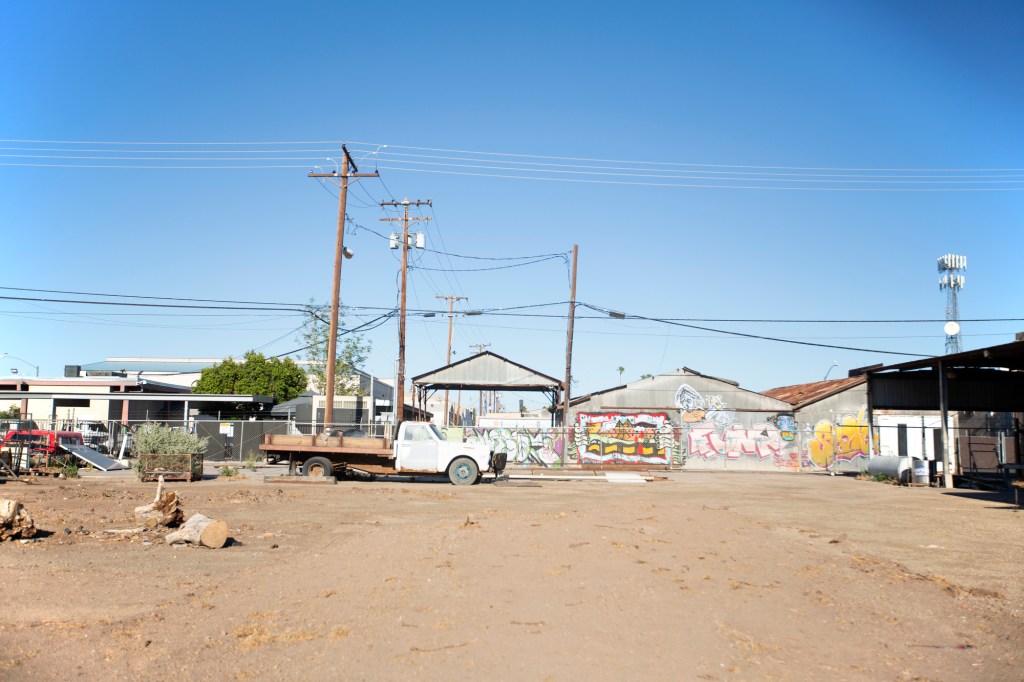 An old truck is parked in an empty and sandy parking lot. Behind it are empty warehouses and telephone poles. This takes place in a rural setting.