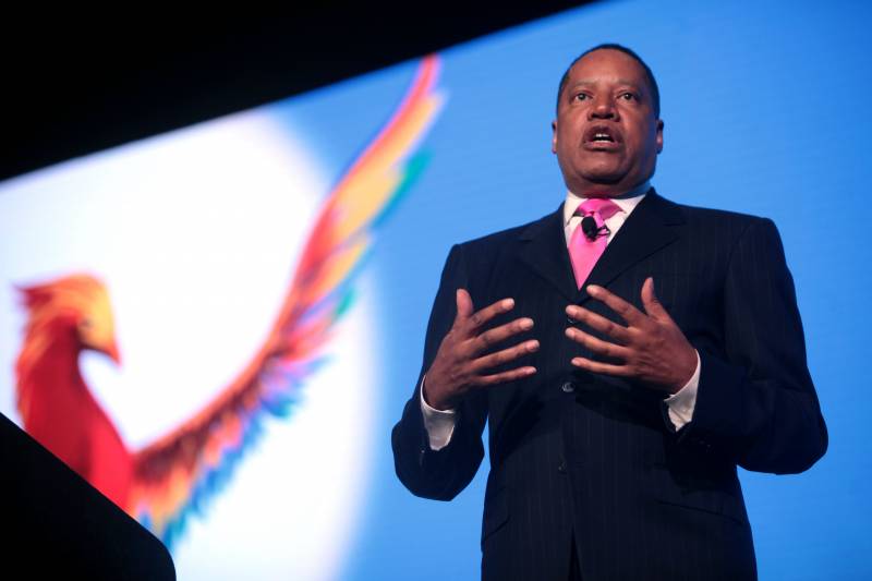 Larry Elder in a dark suit and pink tie, with a projected image of a phoenix on the wall behind him