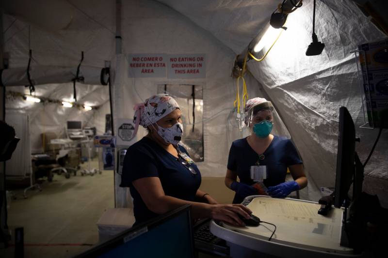 Two nurses wearing scrubs and facemasks look at a screen in a dark tent.