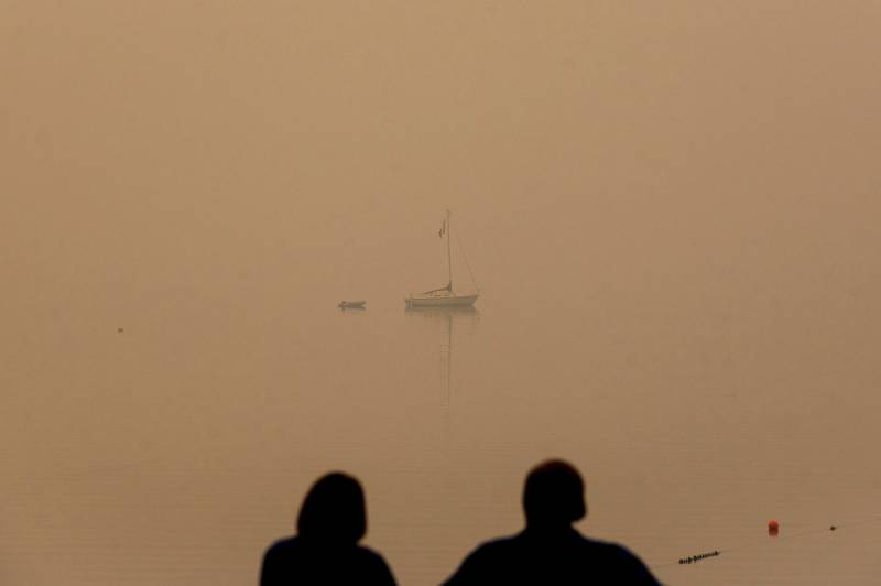 A couple watches from the shore a distant boat on the lake, which has been completely blanketed by smoke.