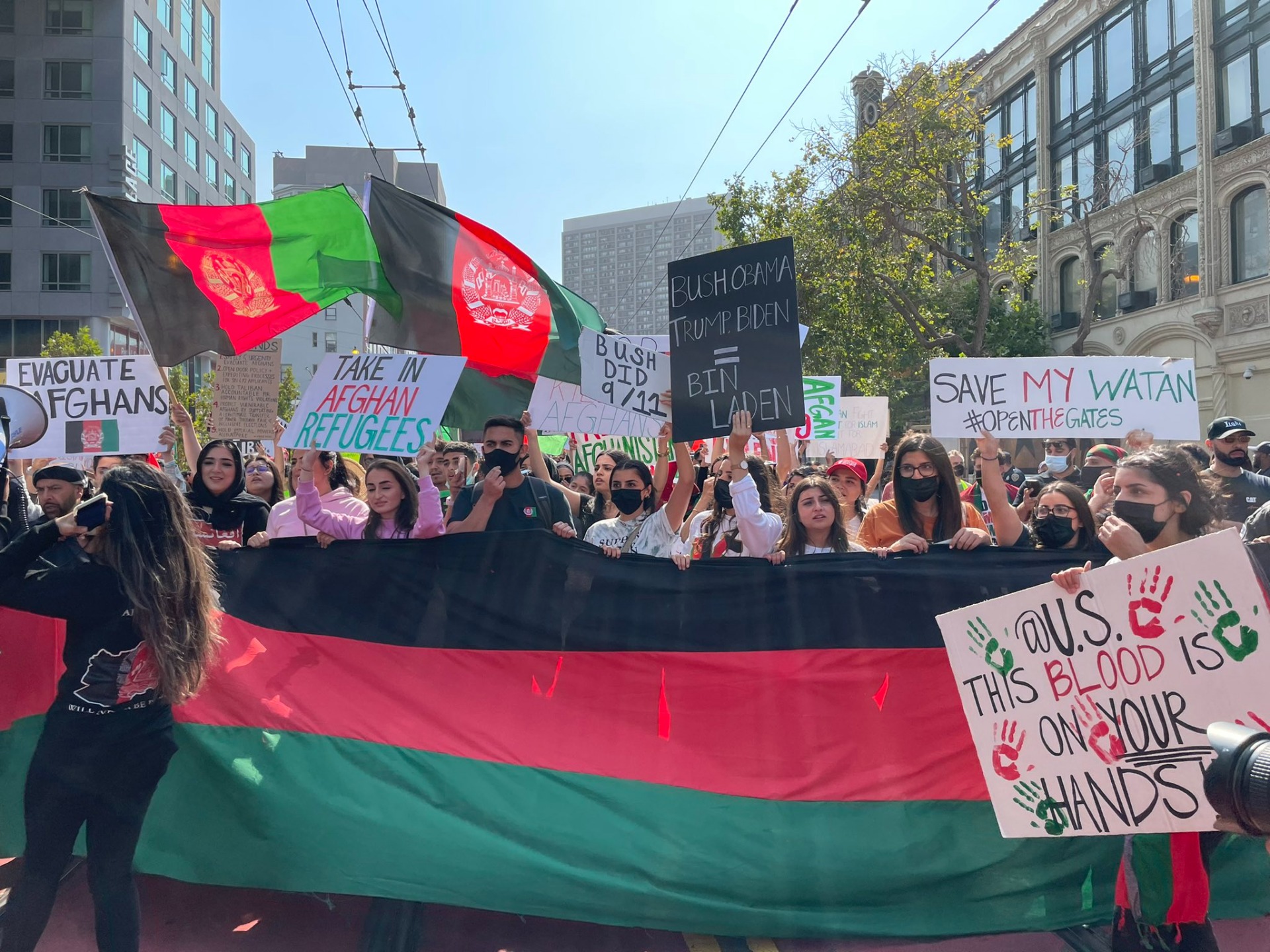 A crowd marching with a long black, red, and green Afghan flag across them, amid protest signs and other Afghan flags