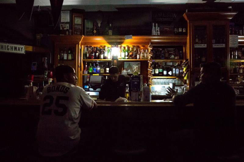 The front side of a bar. The establishment is dark as its lights are not on and only a few lamps provide light to a bartender that is attending to two customers who are talking to one another.