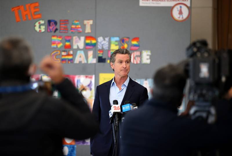 Gavin Newsom, without a tie, speaks into a bank of microphones in front of a board with colorful letters that spell "The Great Kindness Challenge."