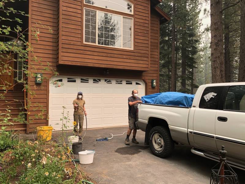 Man and woman packing bed of white pick-up truck in front of two-story house in the woods