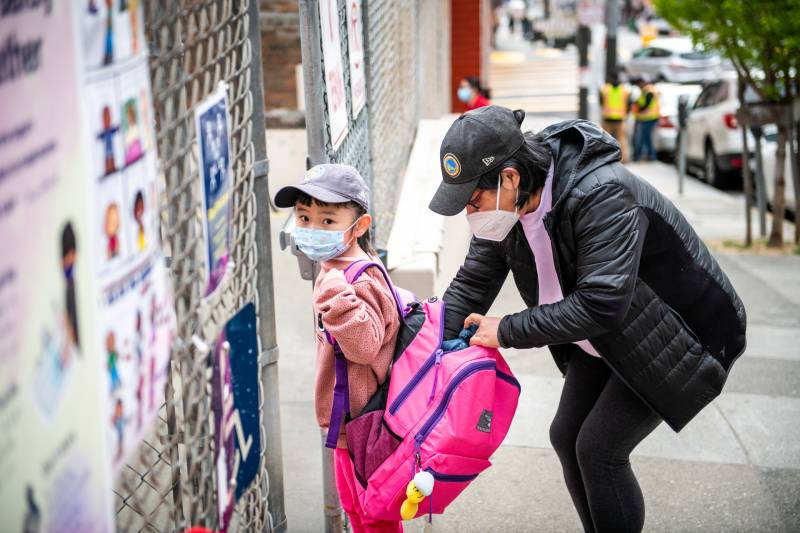 Mom crouches down with hand in young kid's enormous pink backpack, both wearing baseball caps and masks
