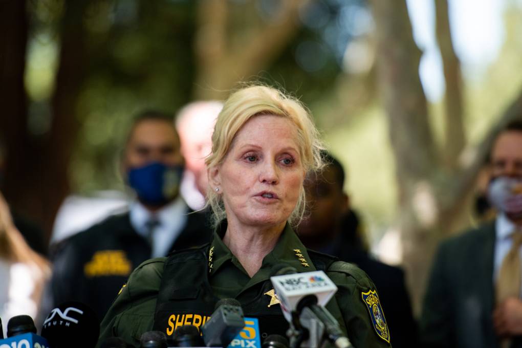 A person with long, blonde hair speaks to a crowd. They are wearing law enforcement uniform and behind them are trees.