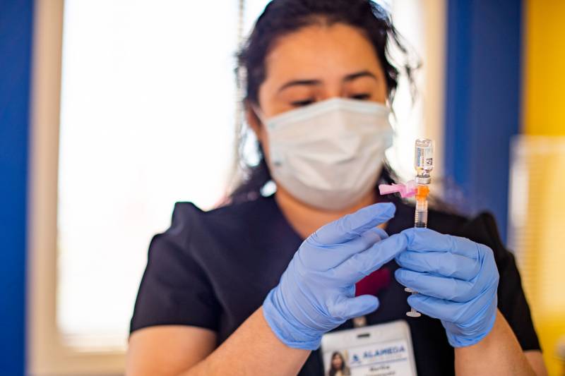 A woman wearing purple gloves flicks a needle inserted upside-down into a vial.