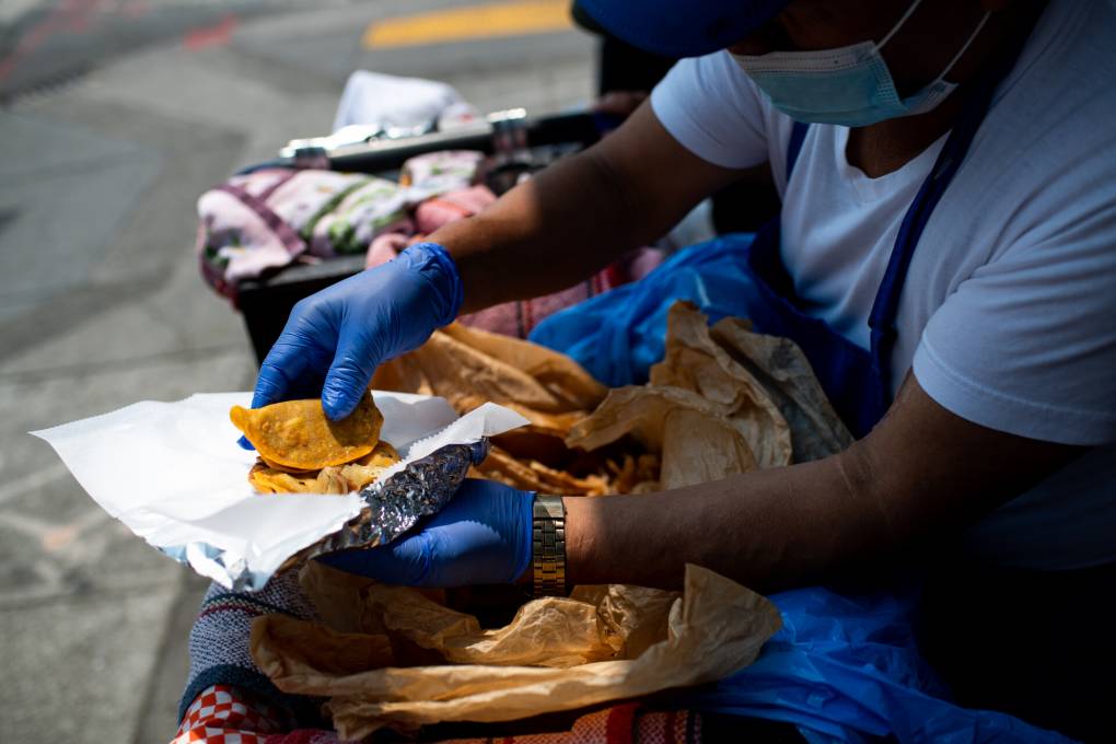 A food vendor stands behind his basket where he keeps his tacos wrapped, wearing gloves and standing in the shade on a sunny day.