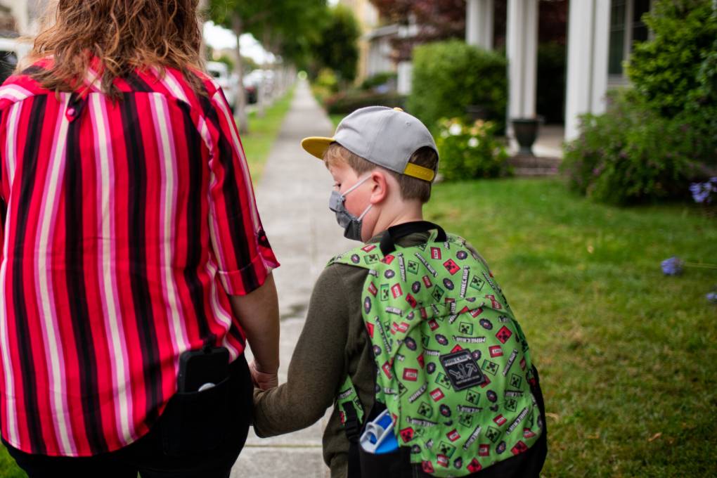 A parent and a child walk on a sidewalk, facing away from the camera. The parent is wearing a red blouse with black and white stripes and the child is wearing a baseball cap and carries a green backpack.