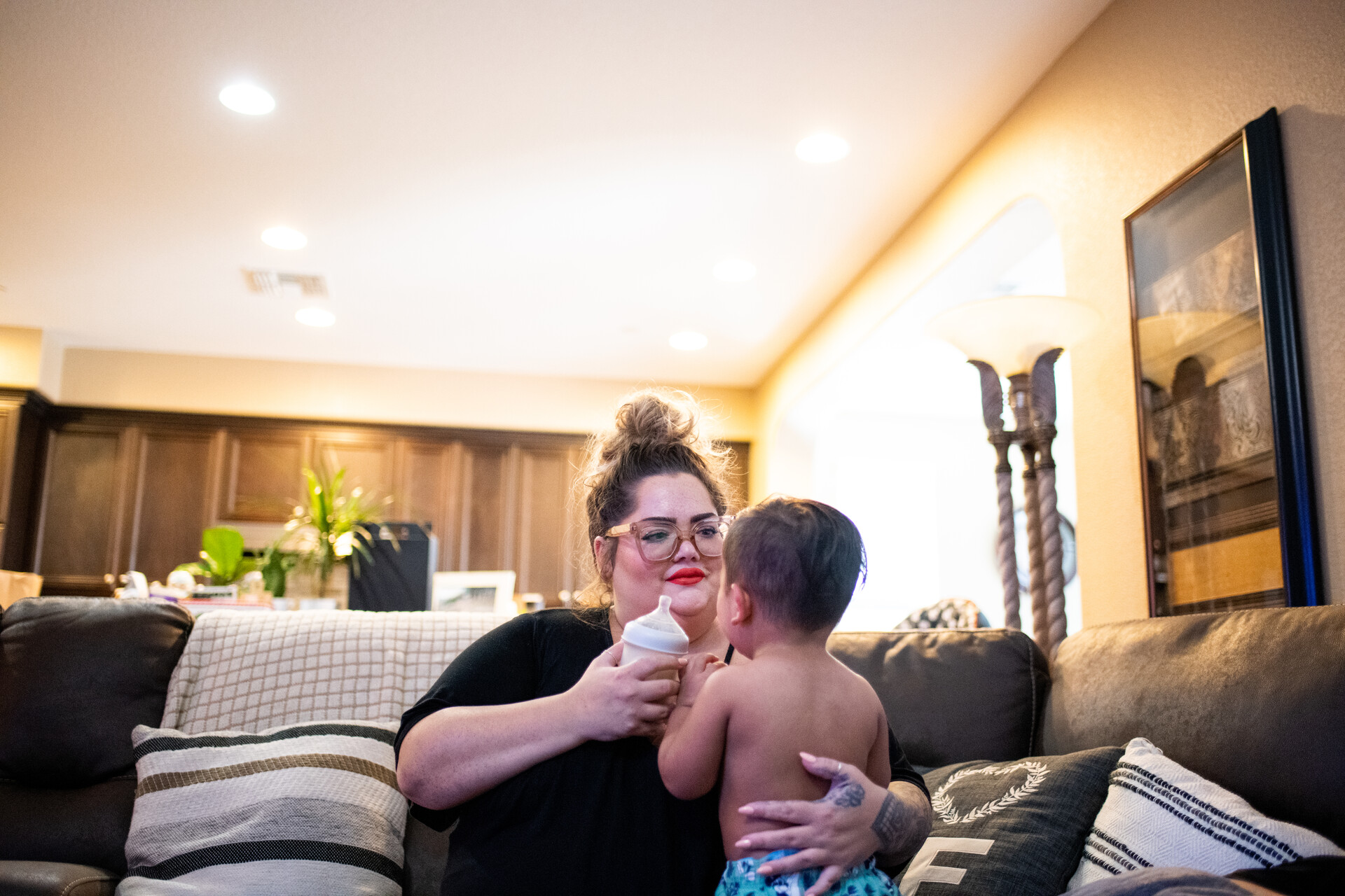 A parent carries a baby in one hand and a baby bottle in the other. The parent is sitting on a big, brown couch in the living room of their house.