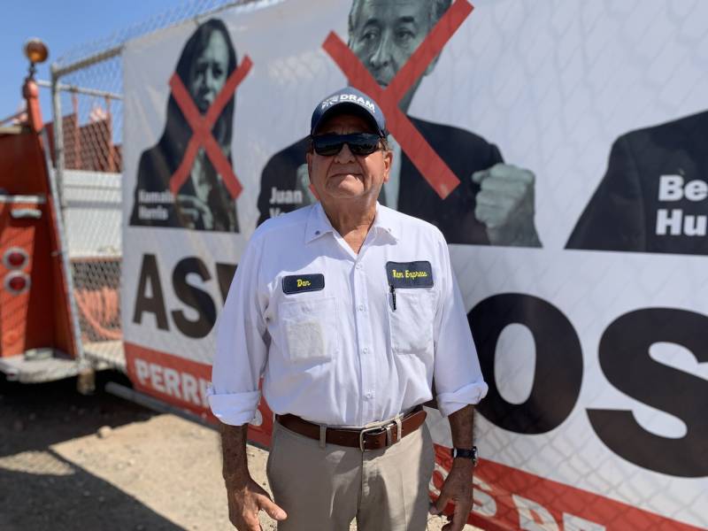 Man in white shirt, sunglasses and blue cap in front of wall-size sign with red Xes over pictures of Kamala Harris and others 