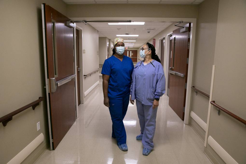 Two nurses dressed in blue scrubs, masks, and blue disposable hospital booties stand in a beige hallway. One looks at the other, one looks at the camera.