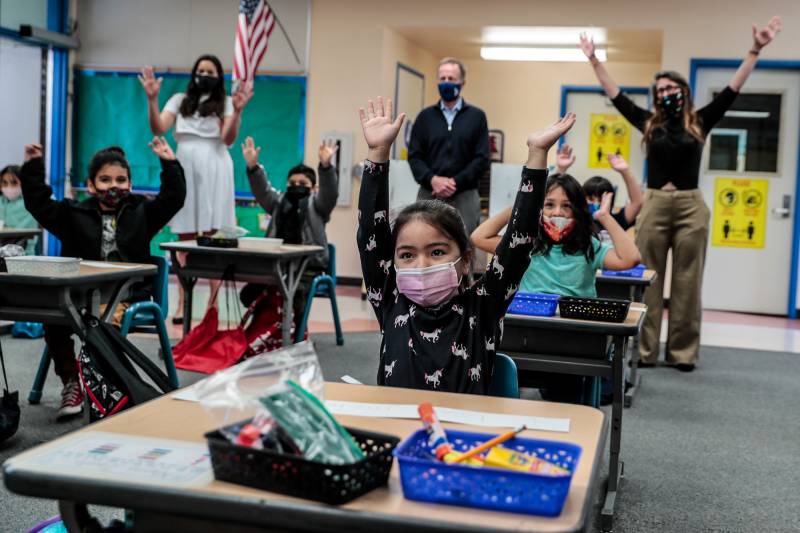 masked kindergartners in masks at desks raising hands