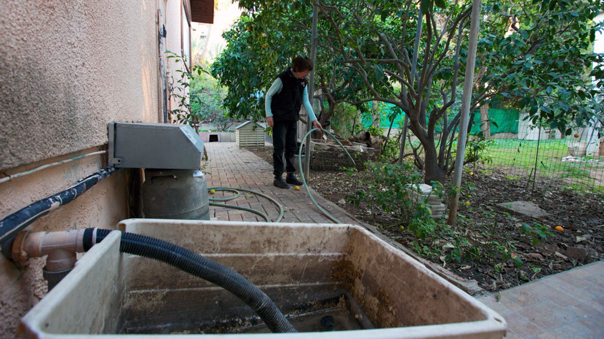 A woman holds a hose connected through a utility sink on the back of her house to water her lawn