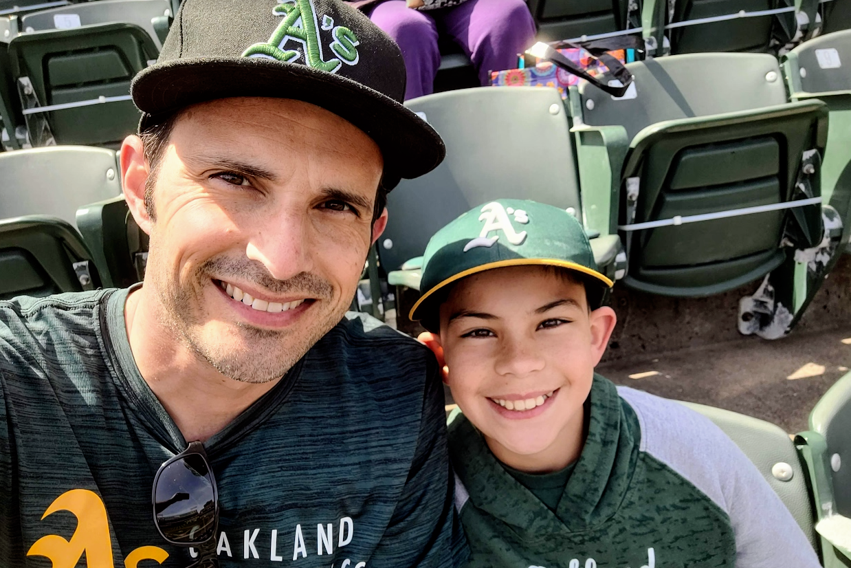 A parent and a child take a selfie. Both are smiling and wear Oakland A's gear.