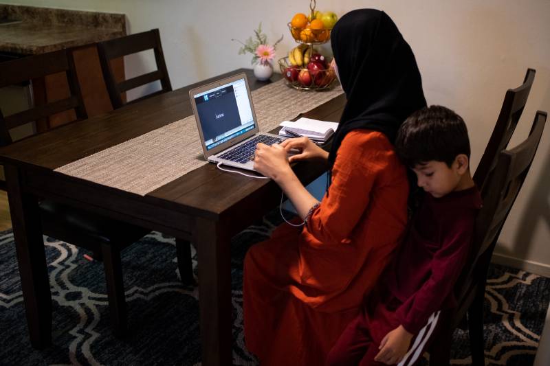 A woman wearing an orange salwar kameez and black hijab sits at a kitchen table typing on a laptop. A boy, age 4, sits right behind her on the chair.