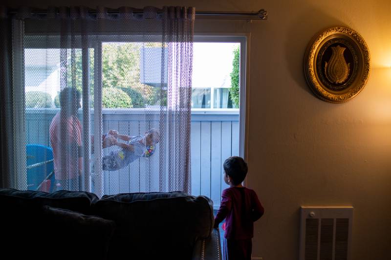 Outside a curtained sliding glass door, a man holds a 2-year-old, as a 4-year-old moves toward the door as if to join them.