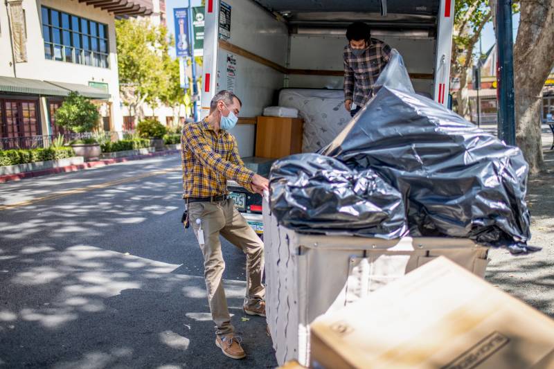 Two men, one inside the back of a truck and one on the street, unload large black garbage bags into a large rolling laundry cart.