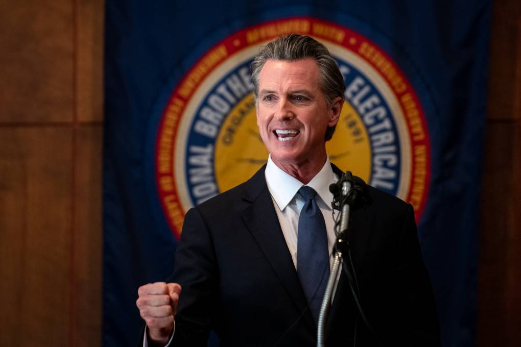 Gov. Newsom speaks flexing his right fist in front of an IBEW union symbol.