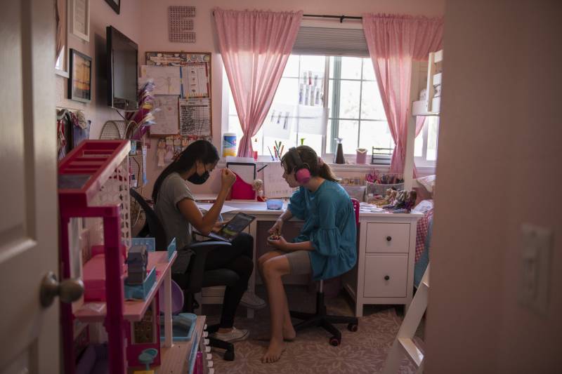 A teenage girl wearing large pink headphones sits in a pink bedroom with a woman holding a laptop.