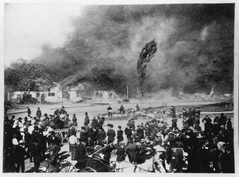 A black-and-white photo of a crowd watching as multiple structures burn down and a large cloud of smoke rises.