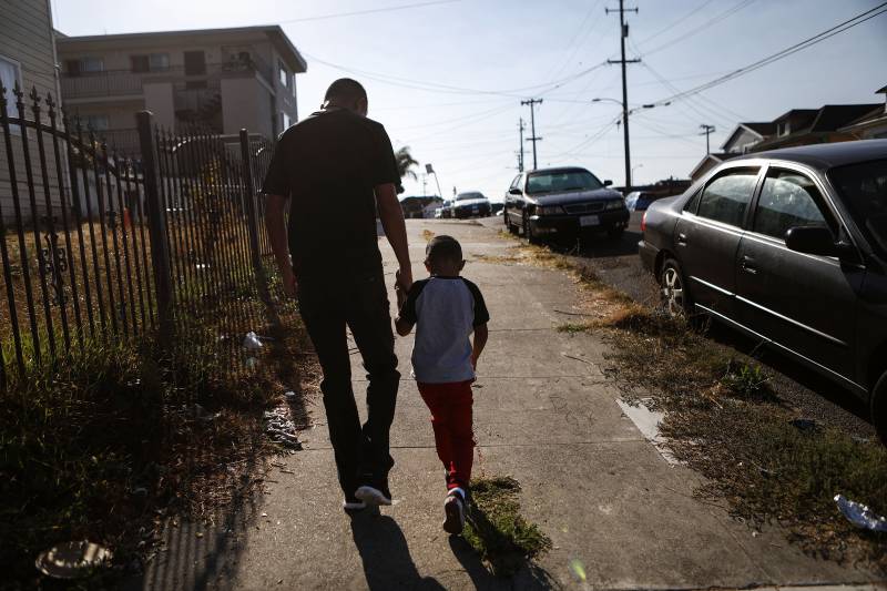 A man and young boy hold hands as they walk in silhouette on an urban sidewalk in early morning sun.