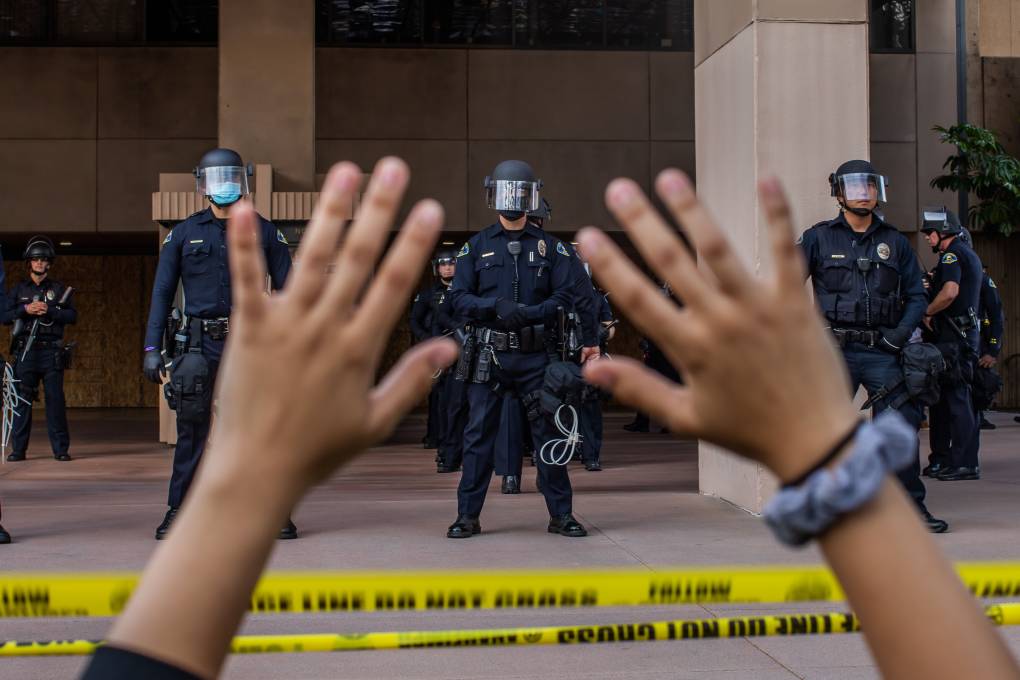 Blurred hands raised in the foreground, with police in battle gear in formation standing on the steps of a building.