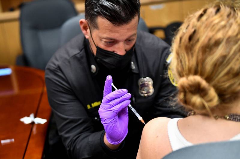 A paramedic wearing a purple glove administers the vaccine to the left arm of a seated person.