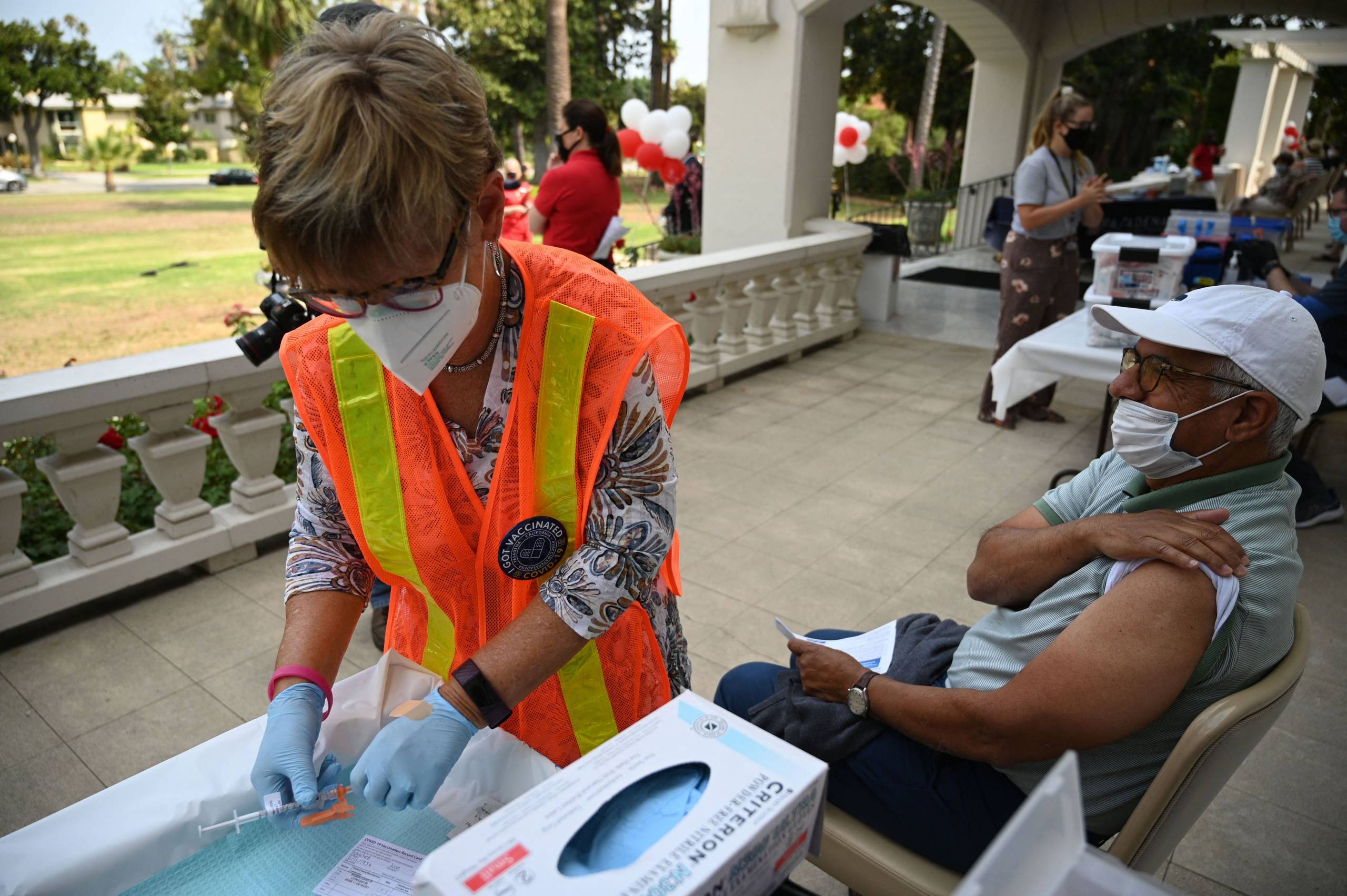 A man sitting on a large porch lifts up his sleeve as he awaits his vaccine, beside a woman in an orange safety vest preparing the vaccine.