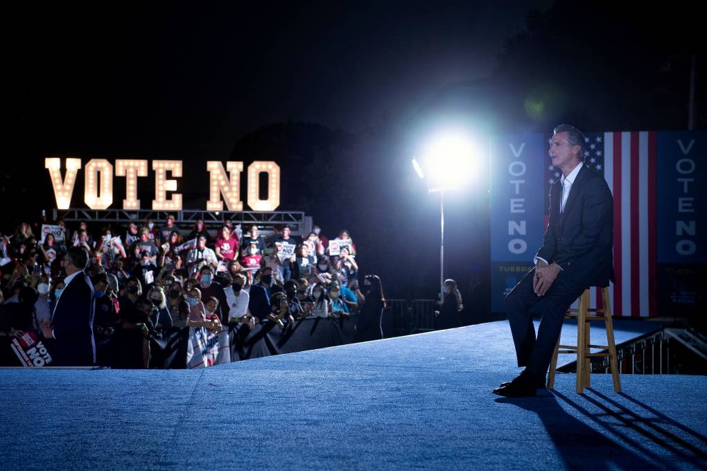 Gov. Gavin Newsom sits on a stool and behind him is a crowd at his rally, also visible is a "Vote No" sign.