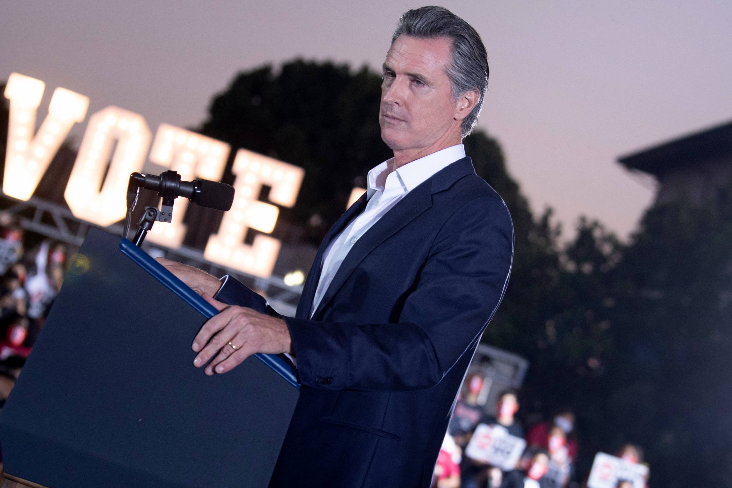 Gov. Gavin Newsom stands at a podium with a stern look, a large lighted sign beyond him that says, "Vote."