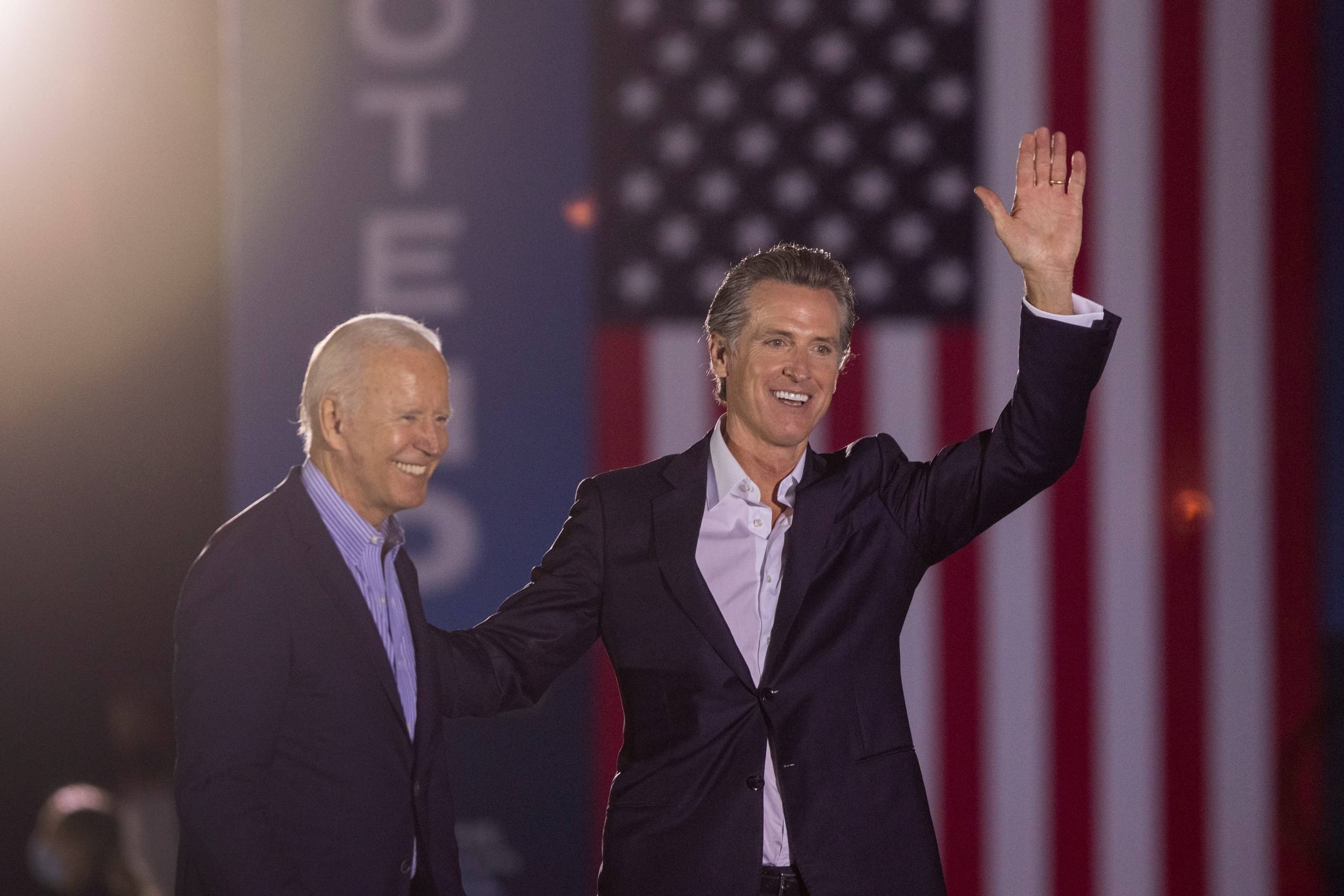 President Joe Biden and Gov. Gavin Newsom, waving, smile as they stand stand before an American flag.