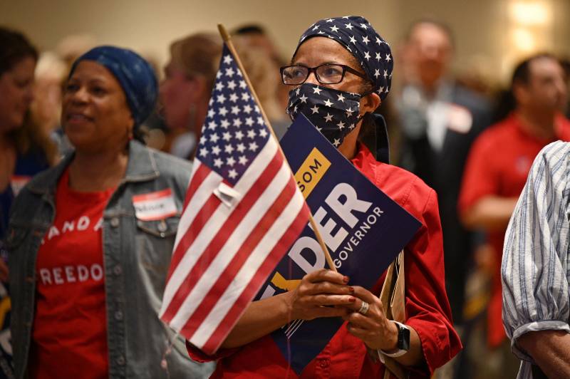 A person clutching an American flag and a Larry Elder poster wears a face mask and bandana printed with the flag.