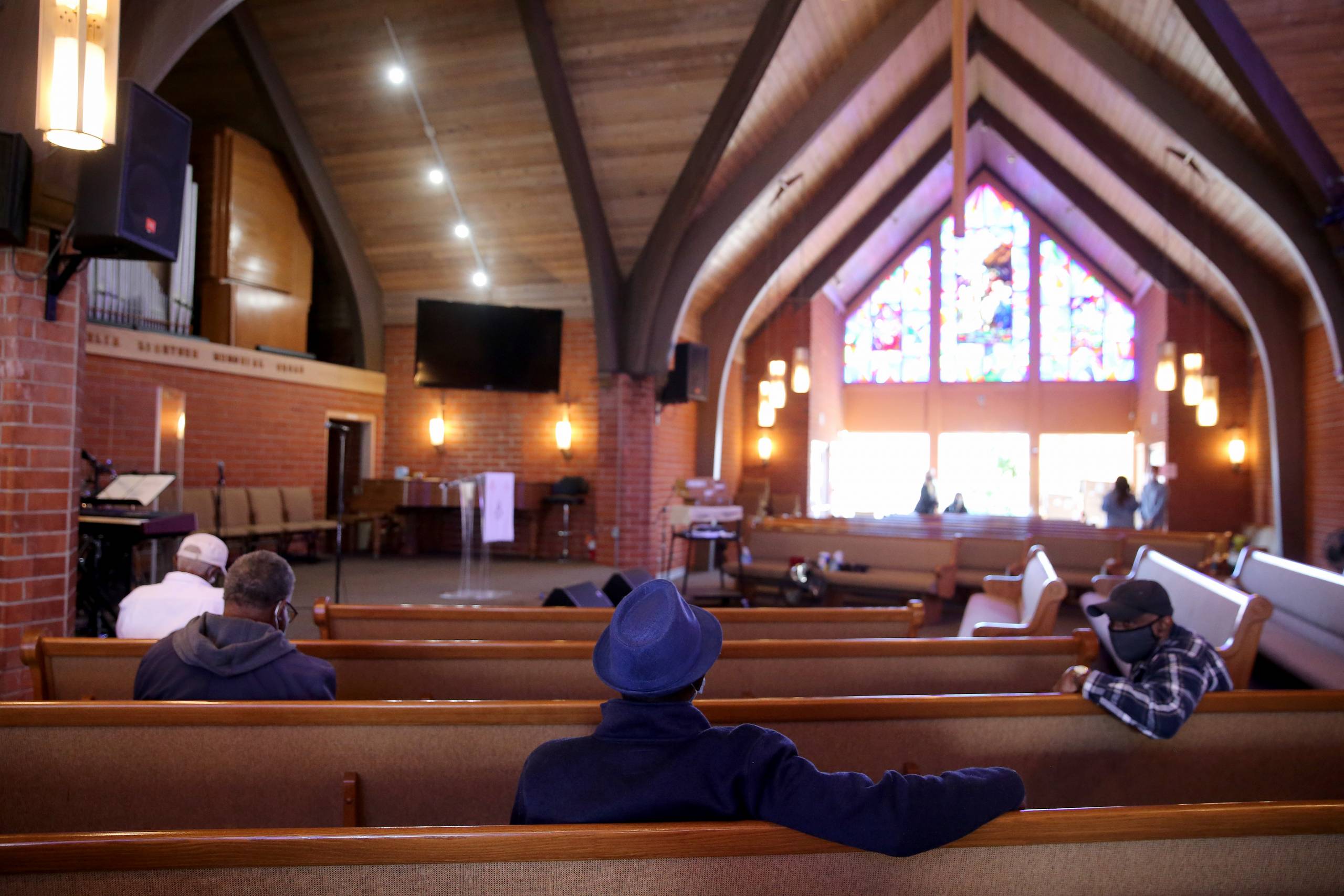 A handful of people sit at a distance from each other in church pews backed by a brightly lit window.