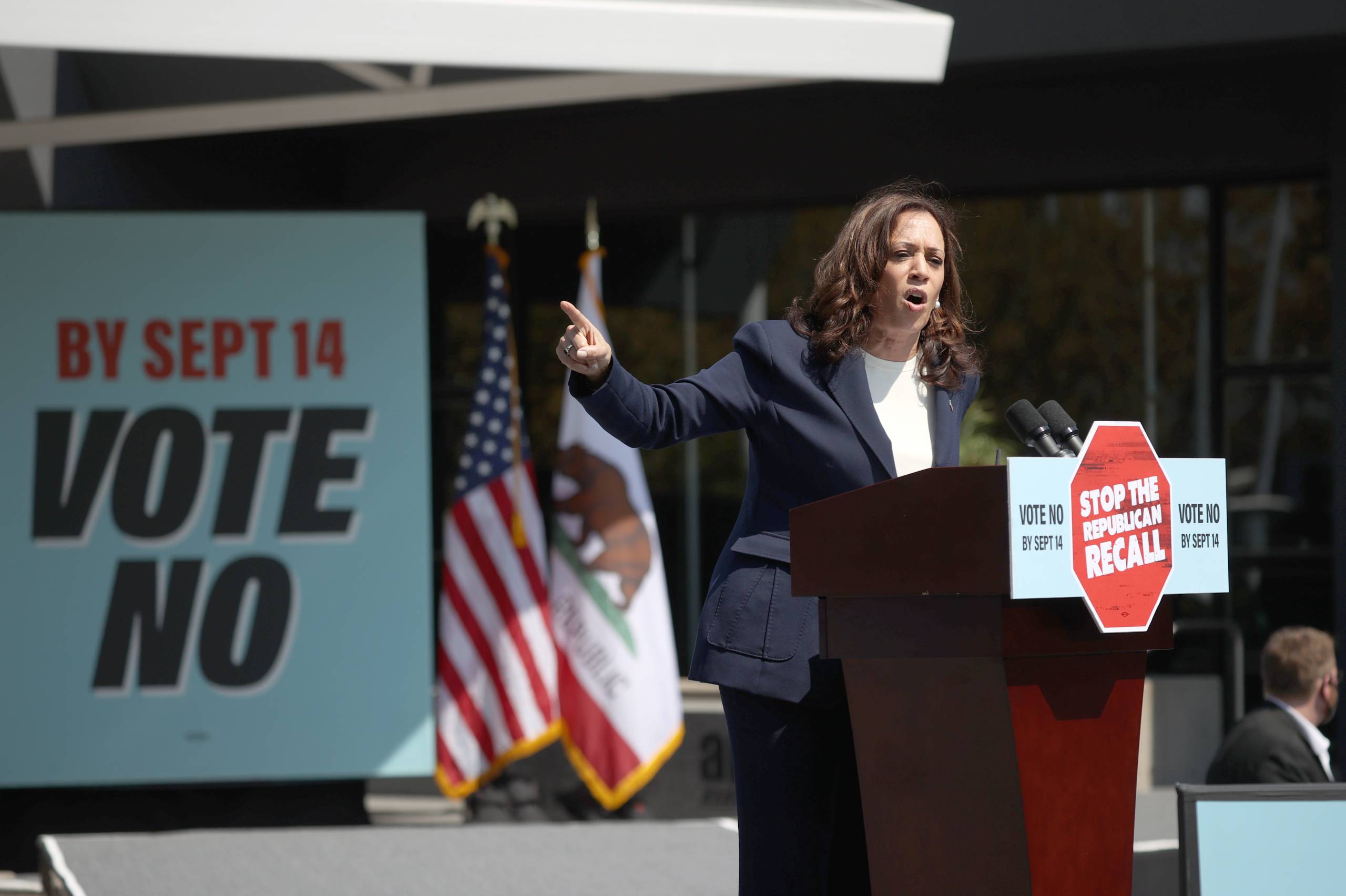 Kamala Harris stands behind a podium and points to her right as she speaks.