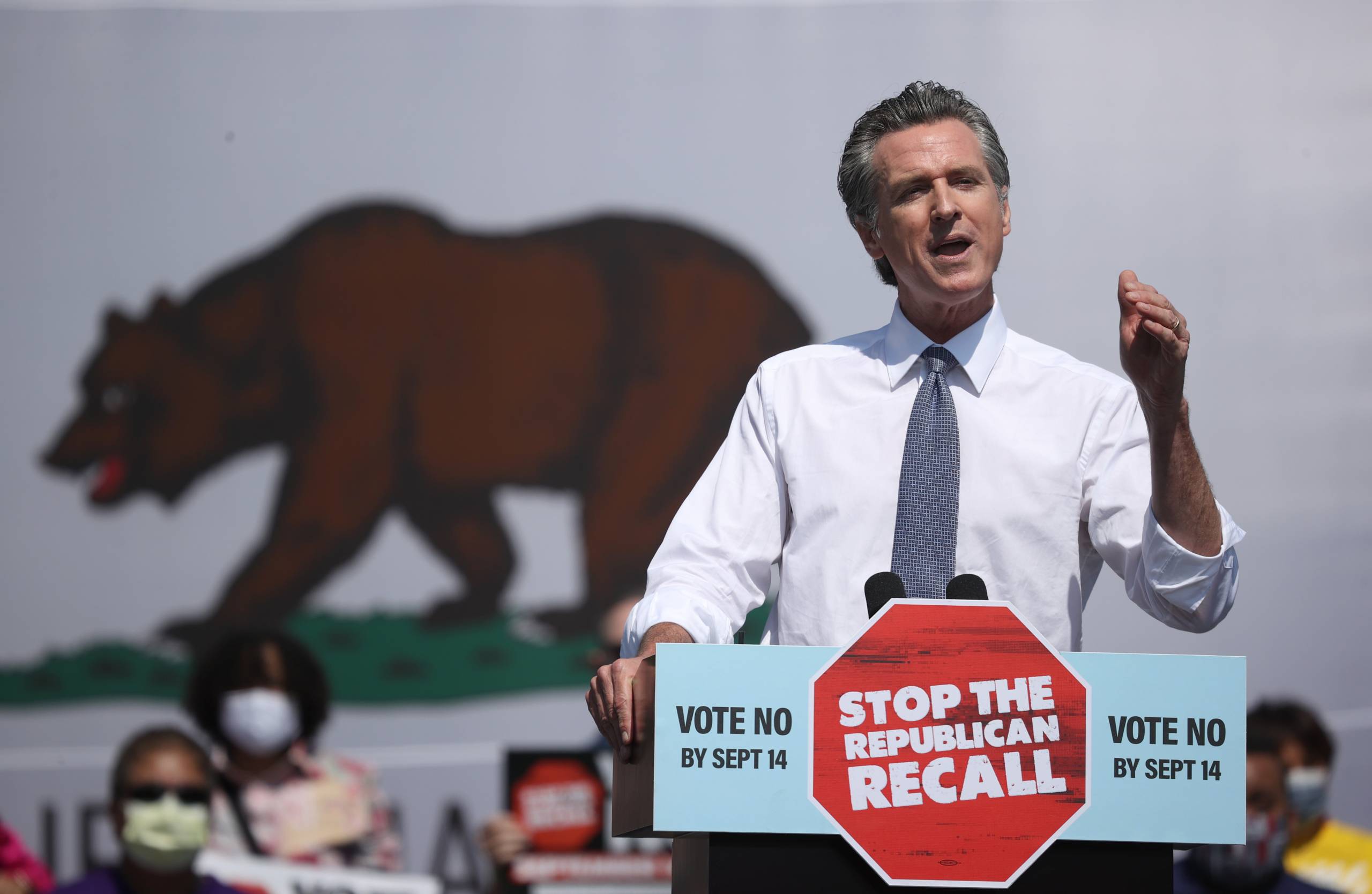 Gov. Newsom gestures behind a podium in front of a massive California flag.