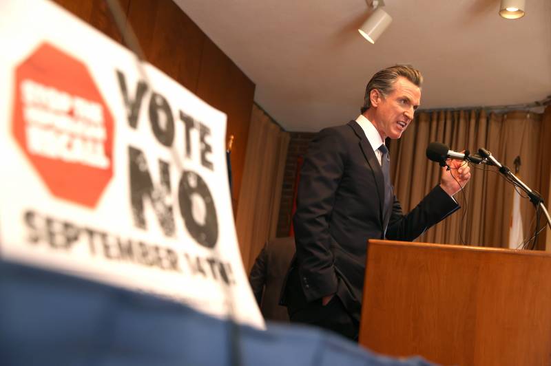 Gov. Newsom brandishes a fist as he speaks at a podium in from a Vote No sign.