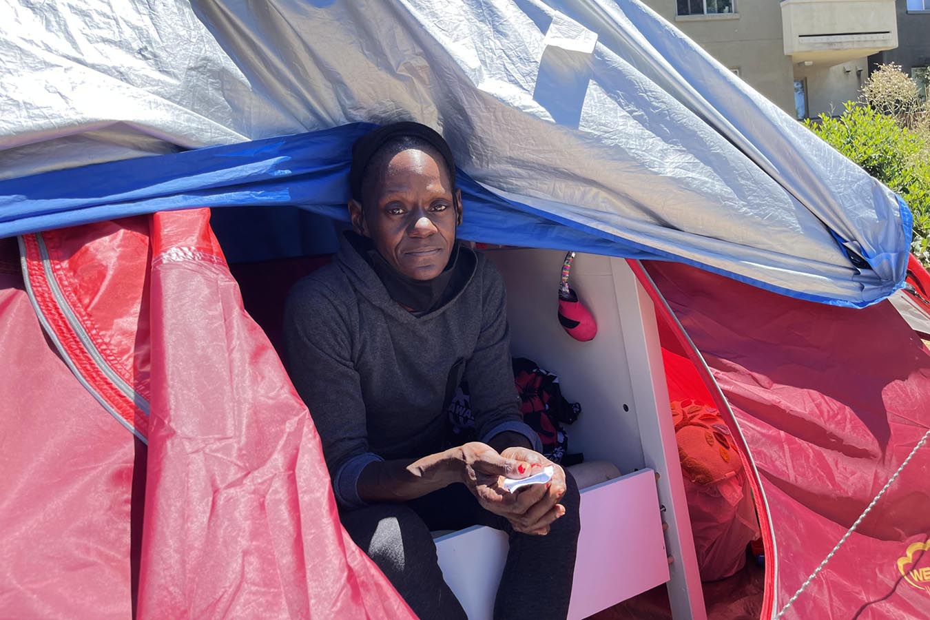 Woman with no hair sits in door of a tent on a bedframe, looking tired.