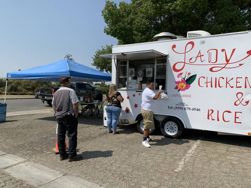 People order food at a white food truck with red lettering and a red flower painted on the side.
