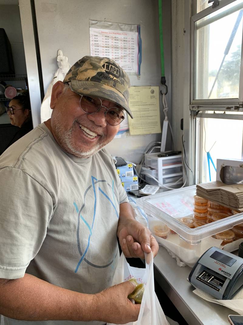 A smiling man in a ball cap and glasses standing inside a food truck puts two small plastic containers into a plastic bag.