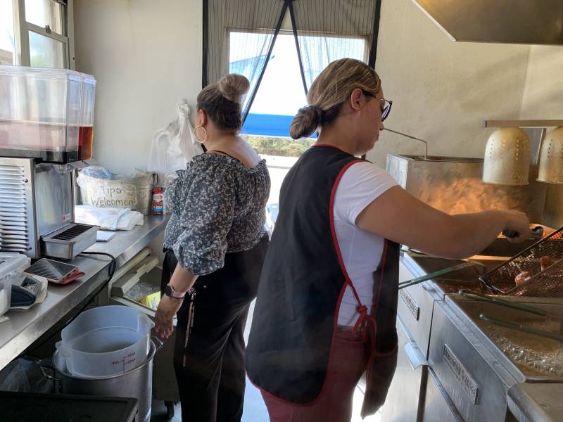 Two women, backs turned, stand inside a food truck, one wearing an apron and pulling chicken from a deep fryer.