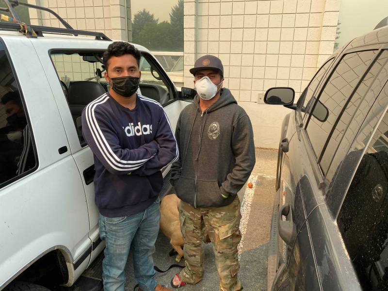 Two men wearing masks and sweatshirts pose between two parked vehicles.
