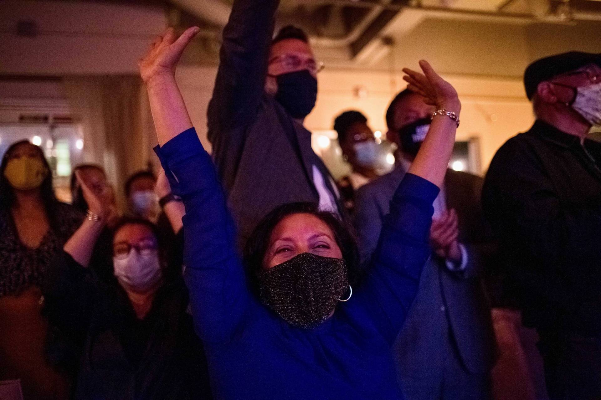 A group of masked Newsom supporters cheer and throw their hands in the air.