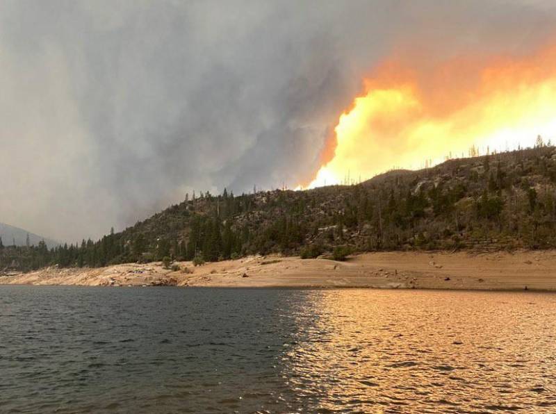 A body of water in the foreground alongside a sandy beach and a forested hillside, beyond which the sky is all flames and smoke.