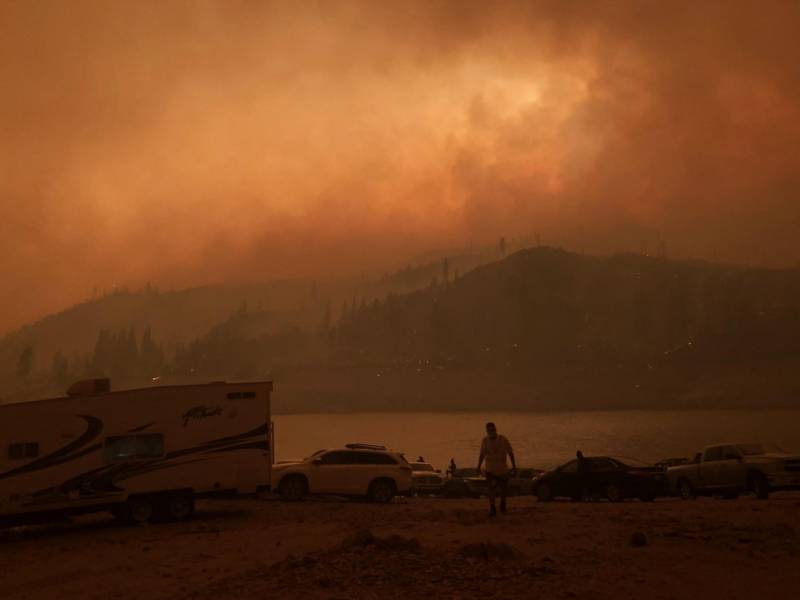 An orange-and-black-hued photo of an RV and a few cars alongside a lake completely darkened by smoky skies.